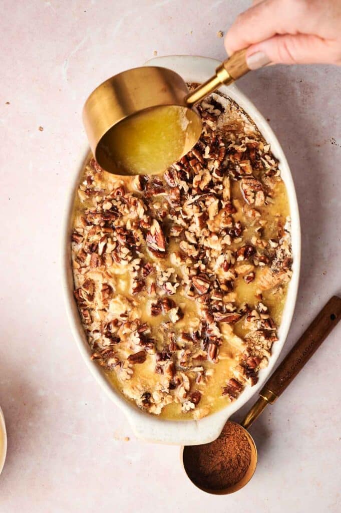 A hand pours melted butter over a dish of pecan-topped sweet potatoes in a white oval baking dish, reminiscent of the rich flavors found in a decadent Pumpkin Dump Cake.