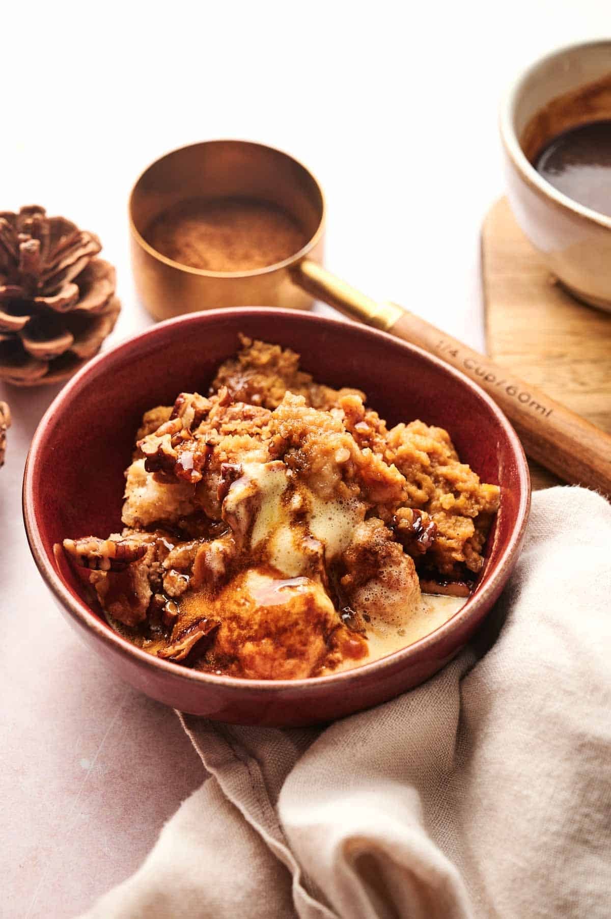 A red bowl of apple crisp topped with melting vanilla ice cream sits next to a measuring cup with cinnamon, a pine cone, and a cloth napkin, offering an autumnal delight reminiscent of the cozy warmth found in every bite of pumpkin dump cake.