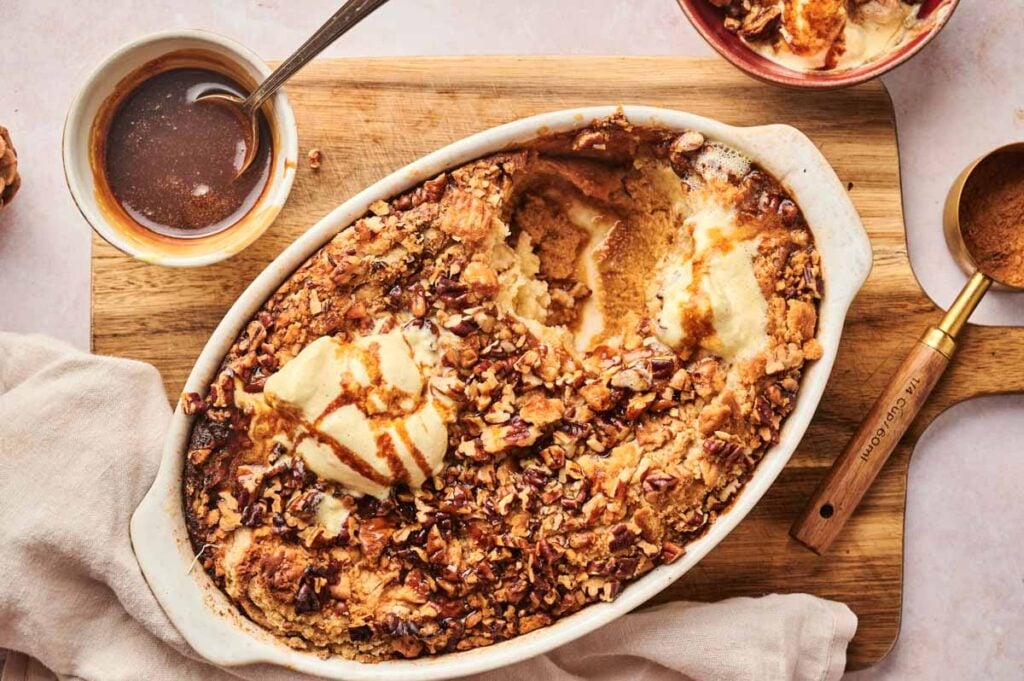 A Pumpkin Dump Cake baked in a white oval dish, topped with vanilla ice cream and chopped pecans, finished with caramel sauce and a bowl of ice cream nearby on a wooden board.