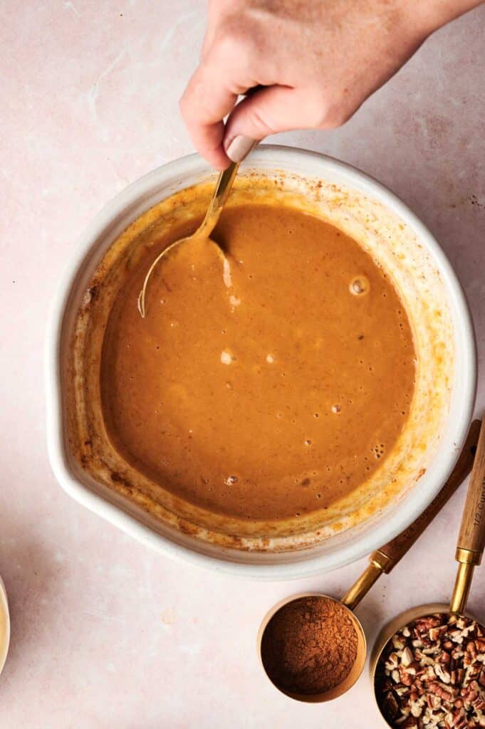 A hand stirs a bowl of pumpkin pie filling with a spoon, setting the stage for a delicious Pumpkin Dump Cake. Nearby, measuring spoons hold cinnamon and chopped nuts, ready to enhance the flavor of this cozy autumn treat.