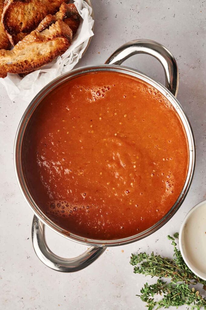 A pot of roasted tomato soup next to a side dish of toast, a small bowl of cream, and a sprig of thyme on a light-colored surface.