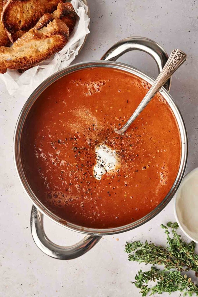 A pot of roasted tomato soup seasoned with black pepper and a dollop of cream, with a spoon in it. Toasted bread and herbs are in the background.