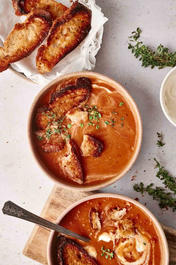 Two bowls of creamy roasted tomato soup topped with toasted bread slices and fresh thyme. A few more toasted bread slices are placed on a sheet of parchment paper beside the bowls.