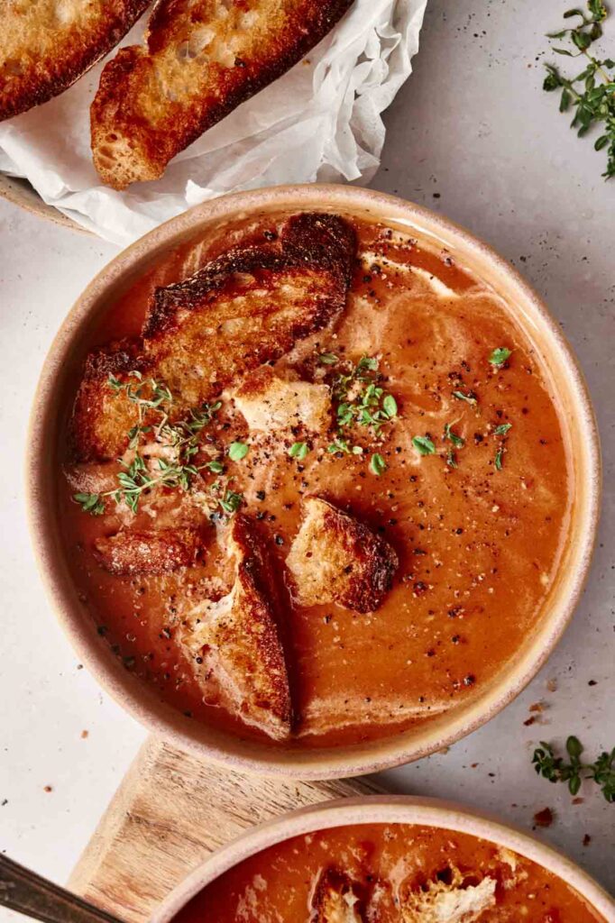 A bowl of roasted tomato soup topped with crispy bread pieces and herbs. A plate with extra pieces of toasted bread is in the background.