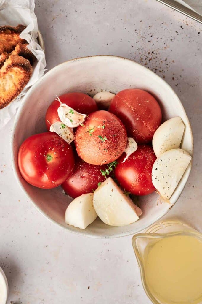 A white bowl containing whole tomatoes, garlic cloves, and onion pieces sits on a countertop, ready to be transformed into a delicious Roasted Tomato Soup. Nearby are toasted slices of bread and a small container of yellow liquid.