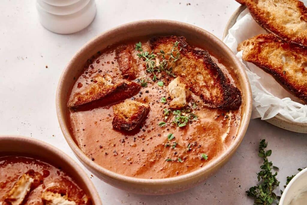 A bowl of creamy roasted tomato soup garnished with croutons and fresh herbs, placed on a light-colored surface with additional croutons on a side plate.