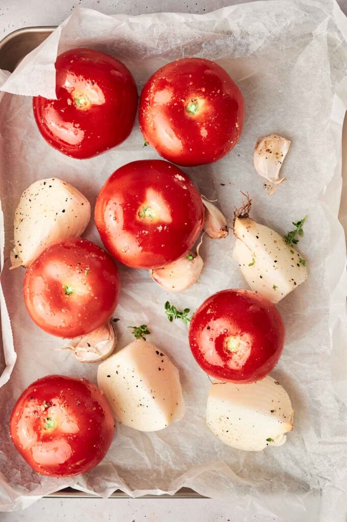 Tray with whole tomatoes, peeled garlic cloves, and halved onions on parchment paper, all sprinkled with salt and pepper—perfect ingredients for a delightful roasted tomato soup.