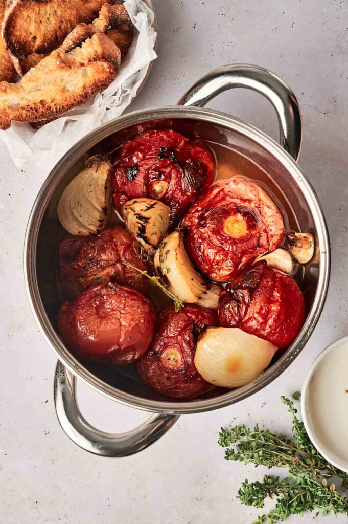 A pot filled with roasted tomatoes and onions on a light countertop, ready to be turned into a delicious roasted tomato soup, with toasted bread on a plate beside it and a sprig of thyme nearby.