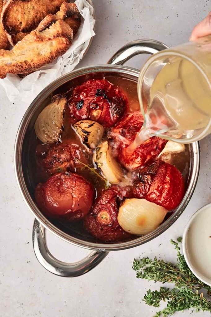 A pot filled with roasted vegetables, including tomatoes, onions, and garlic, having liquid poured into it—a delightful Roasted Tomato Soup in the making. A small plate of toasted bread and some green herbs are placed nearby on a grey surface.