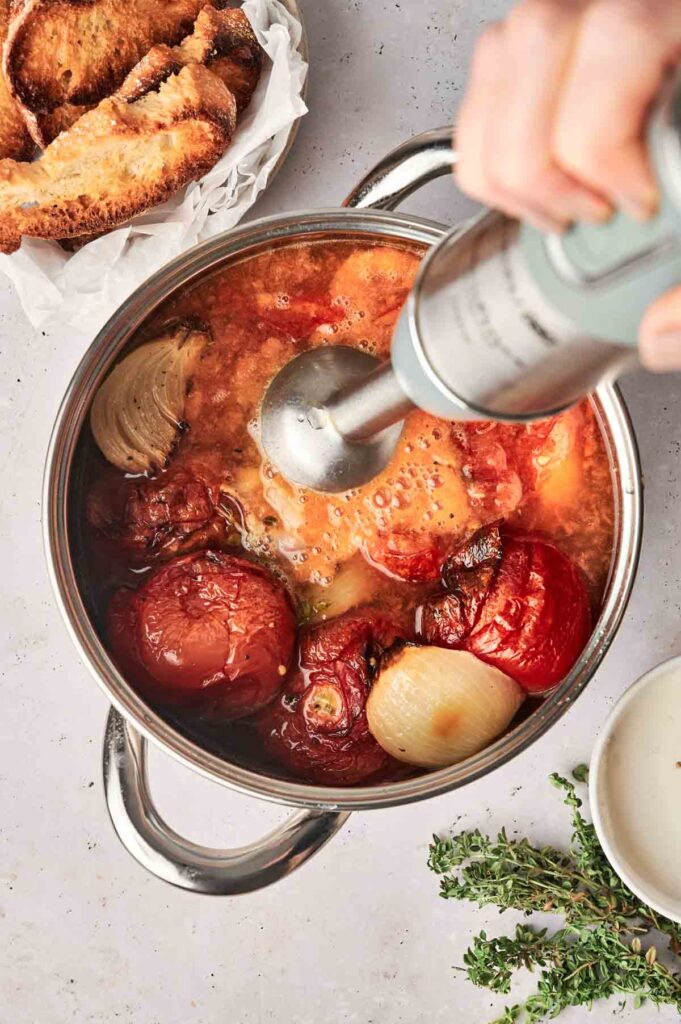 A hand uses an immersion blender to blend roasted tomatoes, onions, and garlic in a pot for a delicious Roasted Tomato Soup. Nearby are toasted bread slices, thyme sprigs, and a small bowl of white sauce.