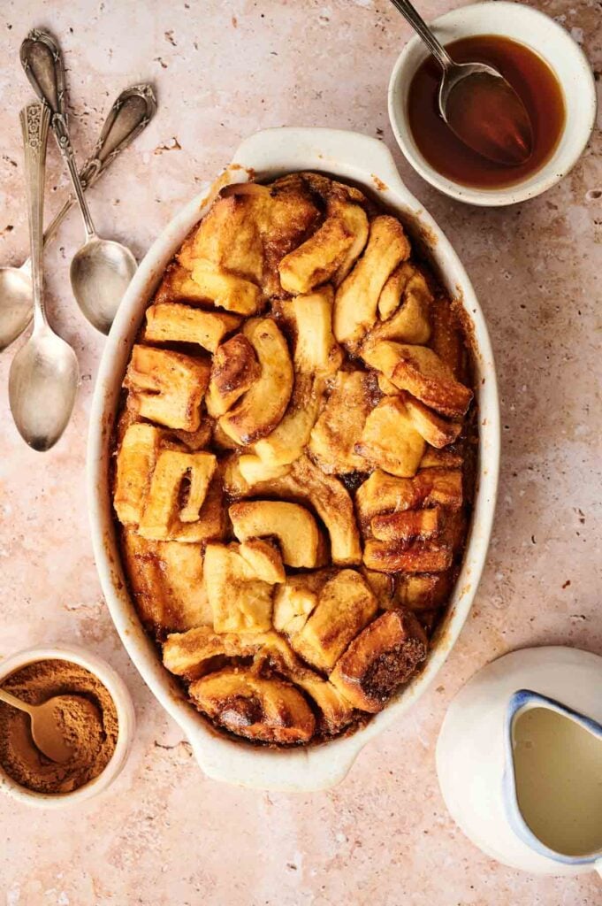 Baked bread pudding in an oval dish with spoons, cinnamon, syrup, and cream on a marble surface.