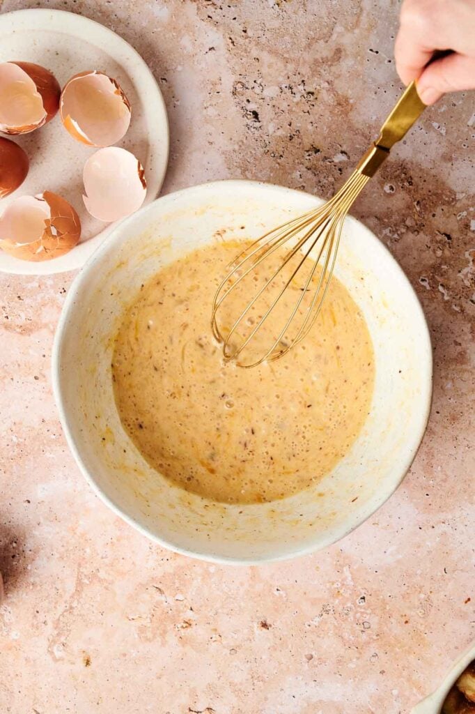 A bowl with mixed batter and a whisk on a countertop, with cracked egg shells on a plate nearby.