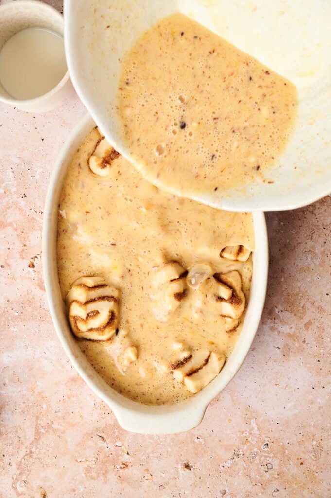 Batter being poured over sliced bananas in a white baking dish on a textured surface.