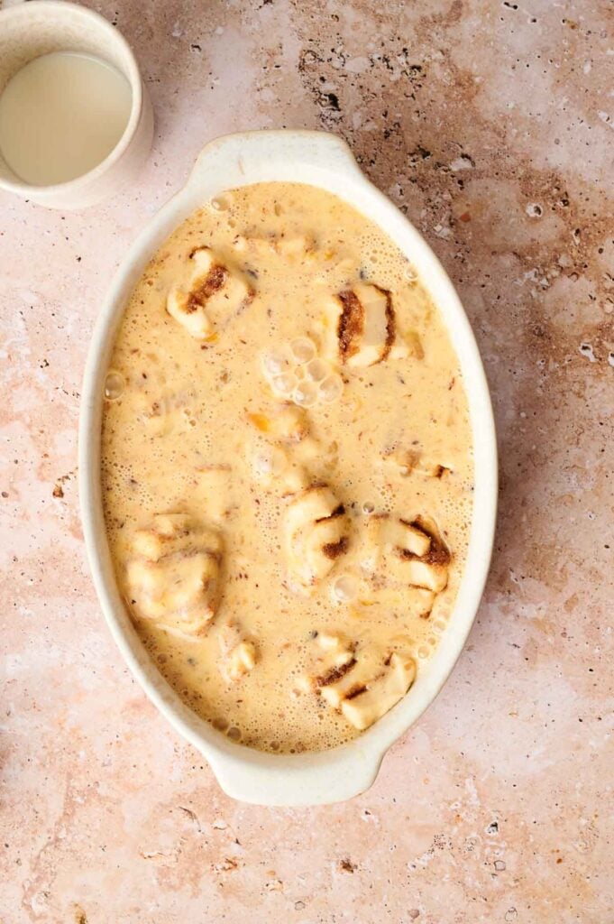 A casserole dish filled with a creamy mixture and pieces of sliced bread is placed on a textured surface next to a small cup of milk.