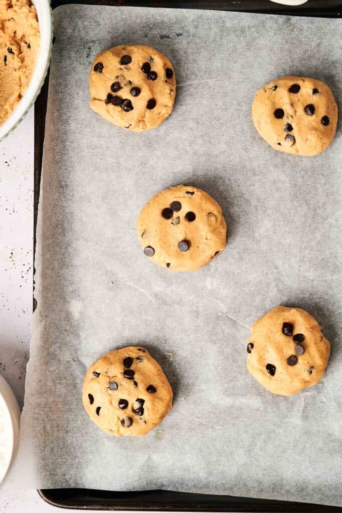 Five unbaked chocolate chip cookie dough balls rest on a parchment-lined baking sheet, perfectly capturing the essence of a copycat Crumbl Cookie recipe.