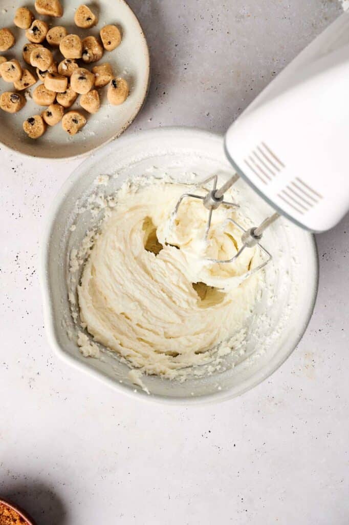 A white mixer is beating dough in a bowl on a light countertop, perfecting the Copycat Crumbl Cookie Dough recipe, with a plate of chocolate chip cookie dough pieces nearby.