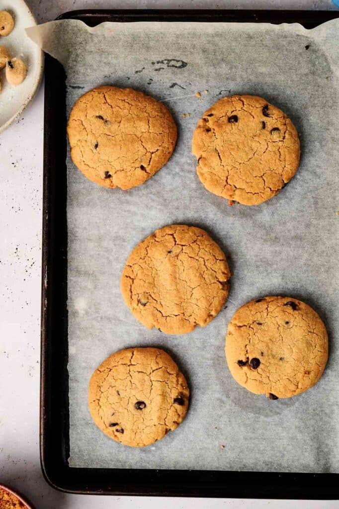 Five chocolate chip cookies rest on parchment paper atop a baking tray, reminiscent of a copycat Crumbl Cookie recipe.