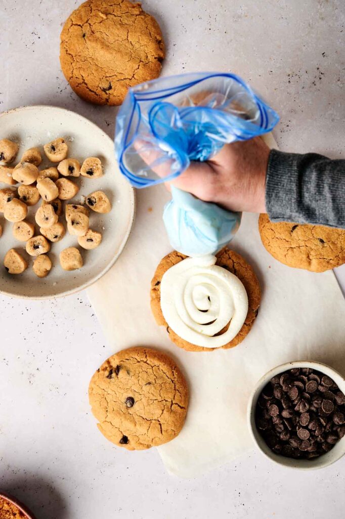 Hand piping cream onto a large cookie, reminiscent of a copycat Crumbl Cookie Dough Cookie recipe, surrounded by more cookies, cookie dough, and a bowl of chocolate chips.