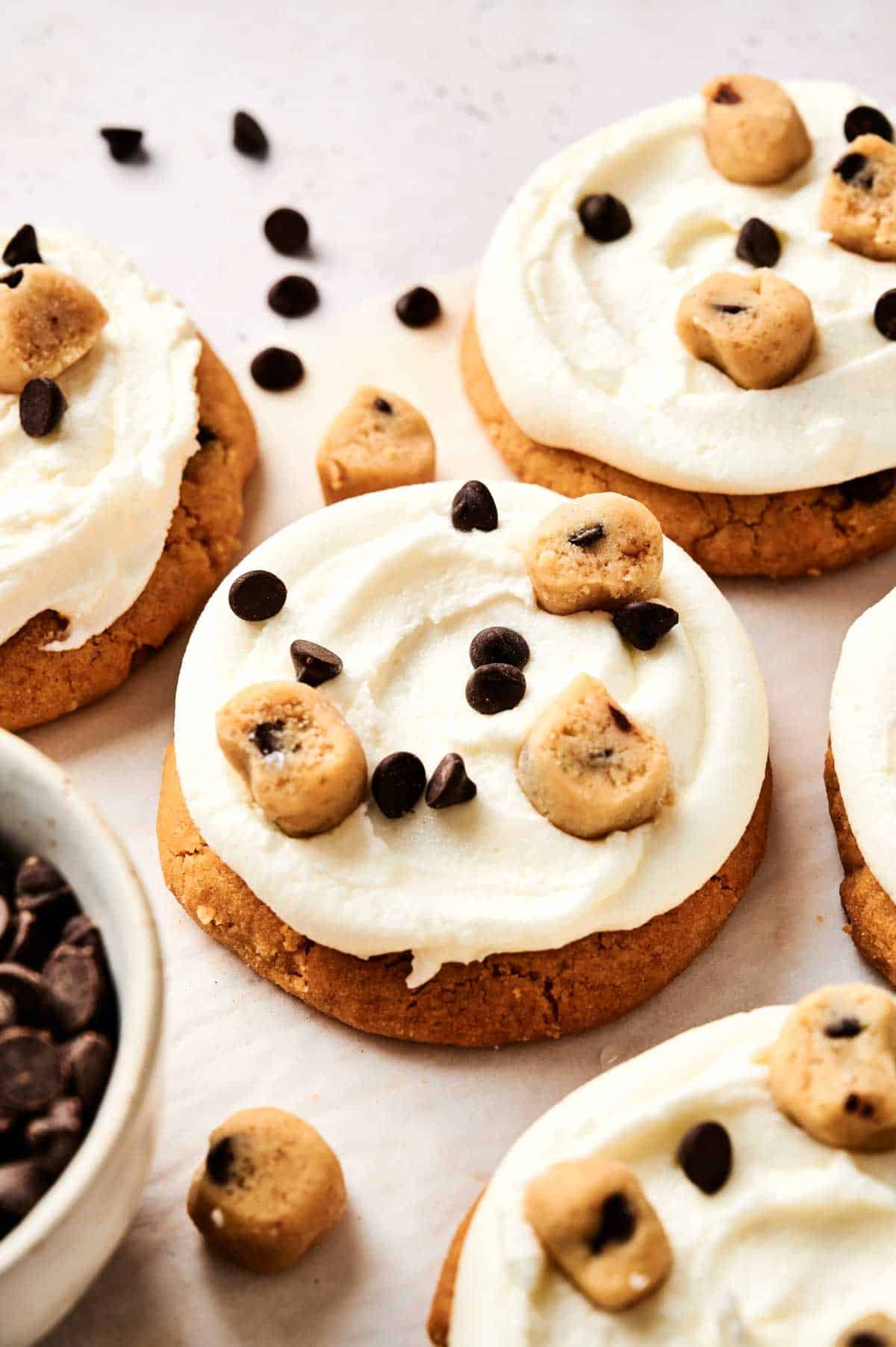 A Copycat Crumbl Cookie Dough Cookie masterpiece awaits: cookies adorned with white frosting, sprinkled with chocolate chips, and crowned with cookie dough pieces, all elegantly arranged on the table. Nearby, a bowl brims with extra chocolate chips for that irresistible touch.