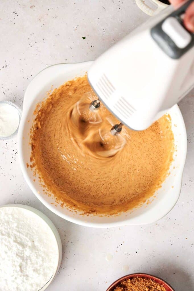 An electric mixer whirs as it blends a brown mixture in a white bowl, surrounded by bowls of flour and brown sugar—perfect for perfecting that Copycat Crumbl Cookie Dough Cookie recipe.