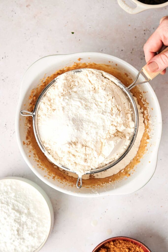 A hand holds a sieve over a mixing bowl to sift flour into a light brown batter for a Copycat Crumbl Cookie Dough Cookie.