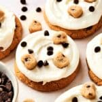 These copycat Crumbl Cookie Dough Cookies feature white frosting, crowned with chocolate chips and cookie dough pieces, on a pristine white surface. A bowl of chocolate chips peeks in from the bottom left corner, inviting you to recreate this delectable recipe.