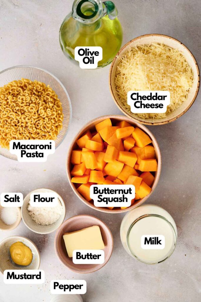 Ingredients for Butternut Squash Mac and Cheese are displayed, featuring macaroni pasta, olive oil, cheddar cheese, creamy butternut squash, milk, butter, flour, mustard, salt, and pepper.