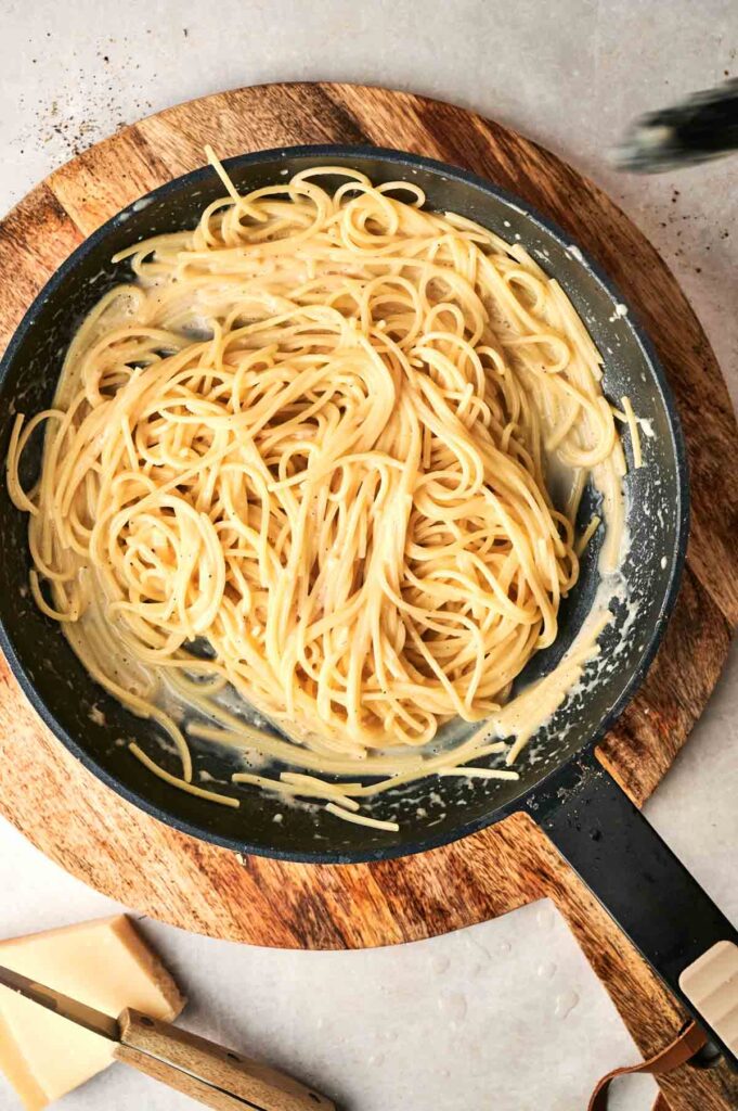 A pan of cooked spaghetti with creamy Cacio e Pepe sauce sits on a wooden cutting board, accompanied by slices of cheese.
