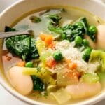A comforting bowl of vegetable soup featuring spinach, peas, carrots, celery, and beans is thoughtfully garnished with grated cheese, with a spoon invitingly placed inside.