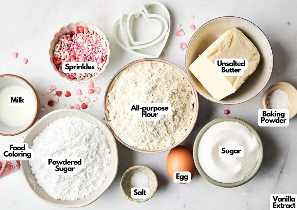 Bowls and cups with labeled ingredients for baking Heart Shaped Cookies: all-purpose flour, unsalted butter, sugar, powdered sugar, baking powder, egg, salt, milk, sprinkles, food coloring, and vanilla extract.