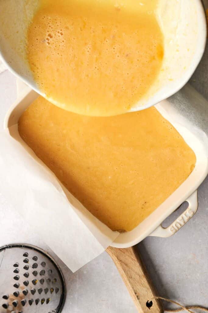 Pouring the zesty lemon bars batter from a bowl into a parchment-lined baking dish on a kitchen counter.