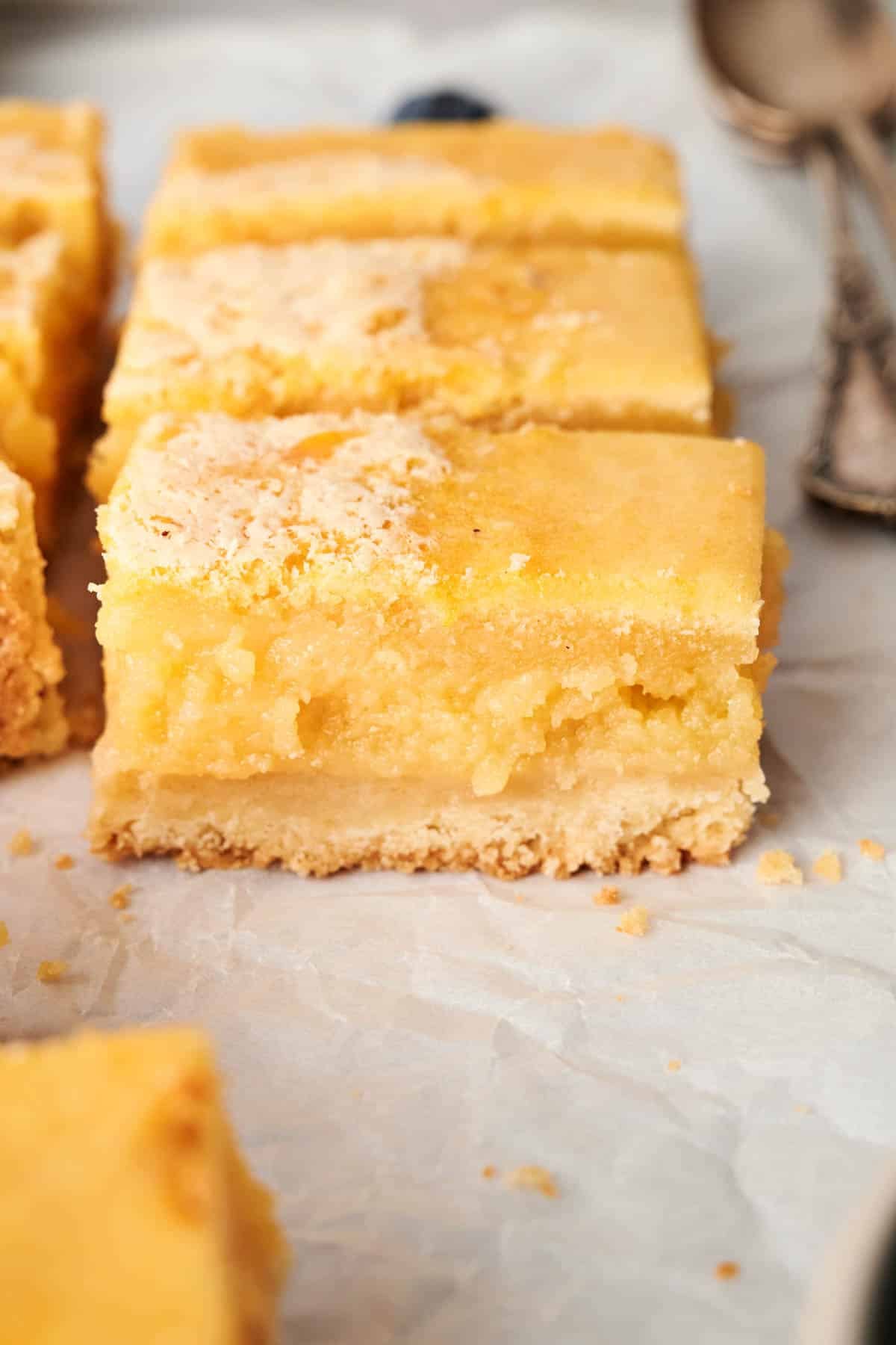 Lemon Bars: A close-up of these tangy delights on parchment paper, showcasing their crumbly shortbread crust and creamy filling. Spoons linger in the background, ready to serve this zesty treat.