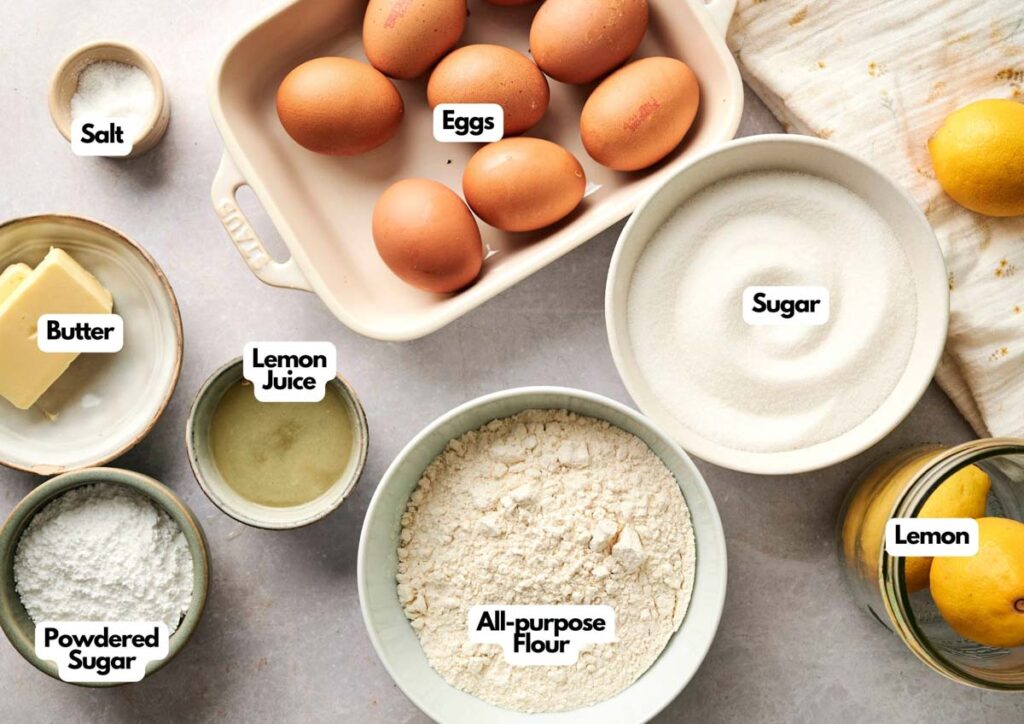 Various baking ingredients for Lemon Bars are displayed on a counter, including eggs, sugar, all-purpose flour, butter, lemon juice, powdered sugar, salt, and a lemon. Each item is labeled for easy reference.