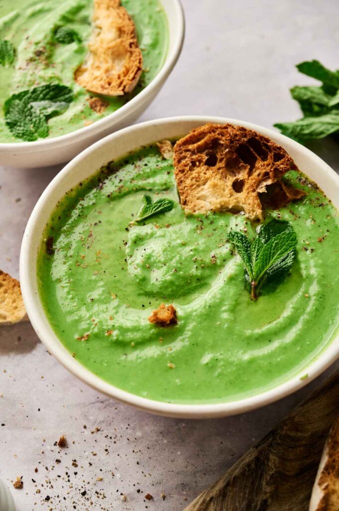 A bowl of vibrant pea and mint soup garnished with fresh mint leaves and crunchy croutons, placed on a light surface.