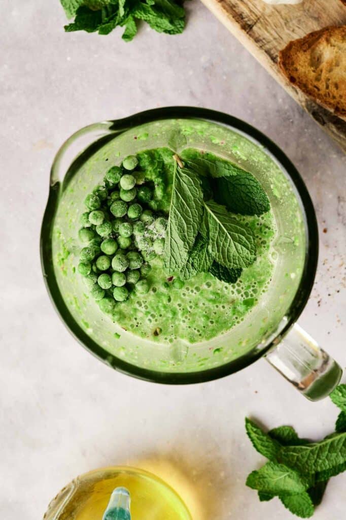 A blender jug brimming with green smoothie ingredients, reminiscent of a refreshing pea and mint soup, sits on a marble countertop. Nearby, fresh mint and slices of bread enhance the scene's culinary charm.