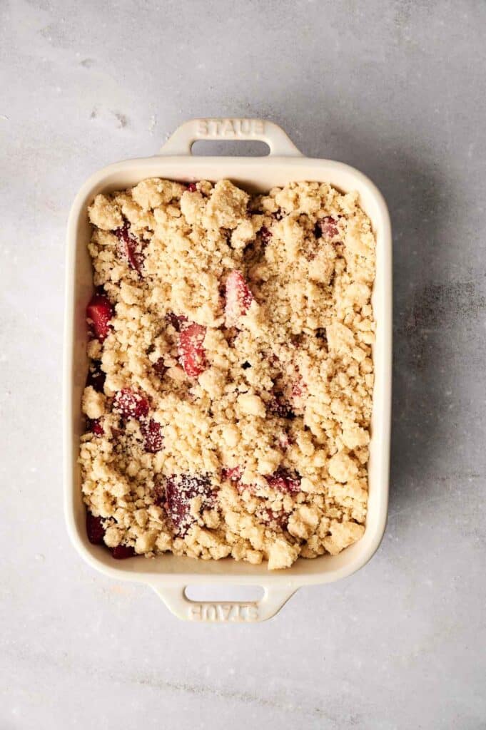 A baking dish brimming with uncooked strawberry dump cake, showcasing a layer of crumbly topping over visible chunks of juicy strawberries, all set against a gray background.