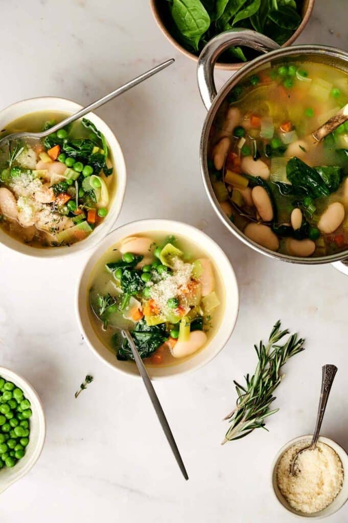A vibrant display of bowls filled with vegetable soup featuring spinach, peas, and beans sits on a pristine white surface. Nearby, you'll find a steaming pot of soup, a bowl brimming with fresh peas, and a small dish offering grated cheese for the perfect finishing touch.