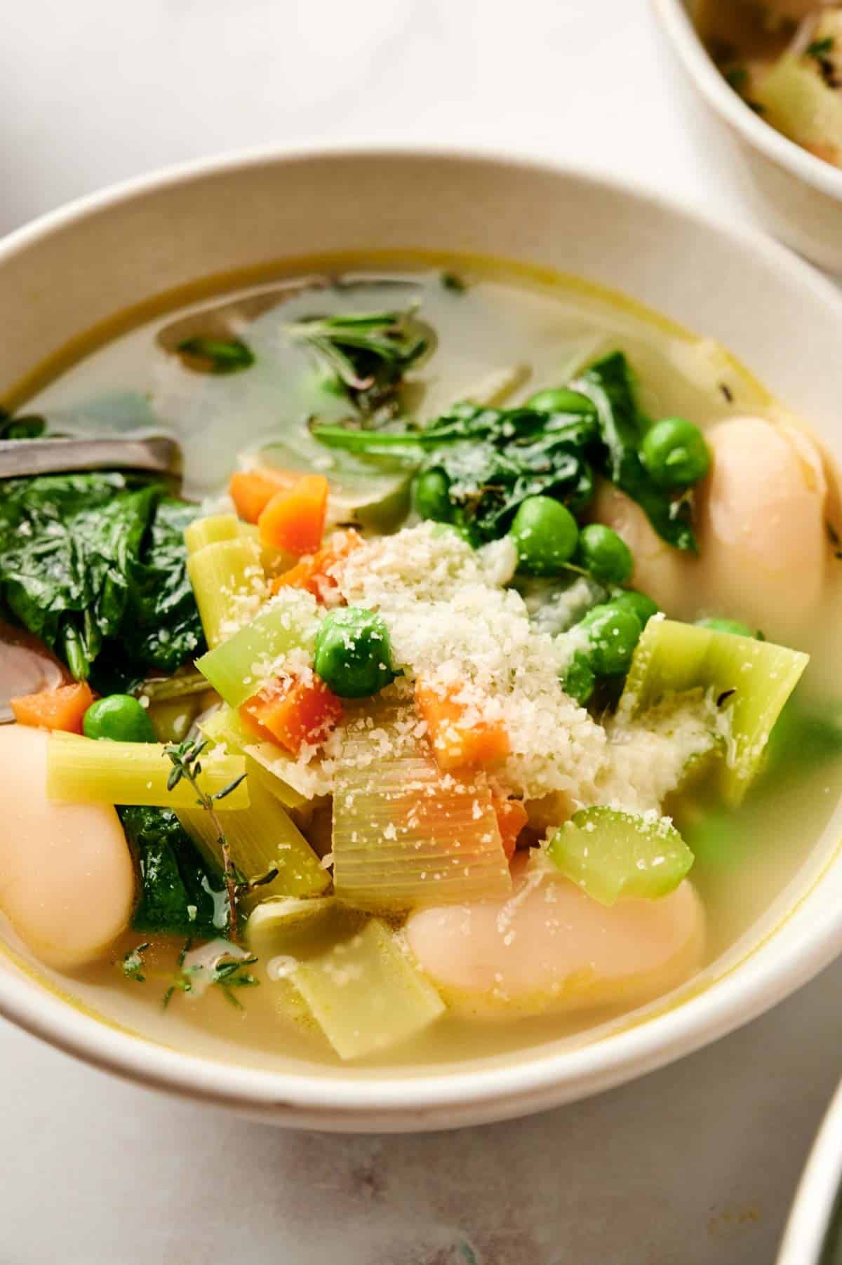A warm bowl of hearty vegetable soup brimming with spinach, peas, celery, carrots, and large white beans, all topped with a sprinkle of grated cheese.