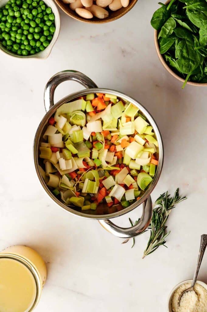 Chopped vegetables, including leeks and carrots, simmer in a pot of bubbling vegetable soup. Surrounding are bowls of peas, beans, and spinach, along with a sprig of rosemary and a small container of grated cheese for added flavor.
