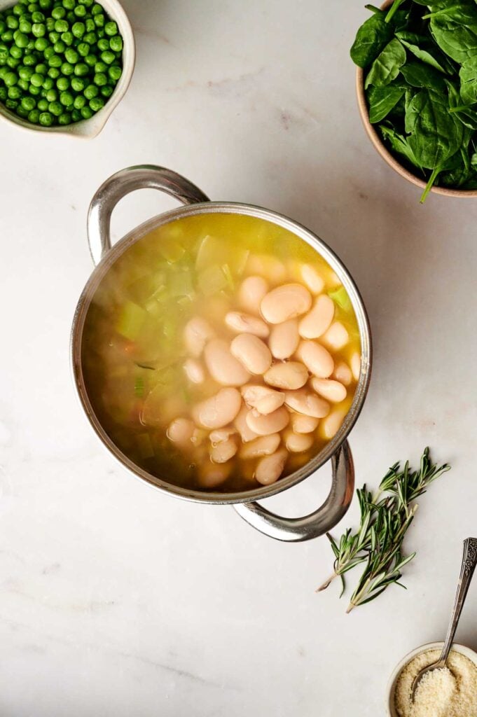 A pot of vegetable soup with white beans and broth sits on a counter, surrounded by bowls of peas, spinach, a spoon, and sprigs of rosemary.