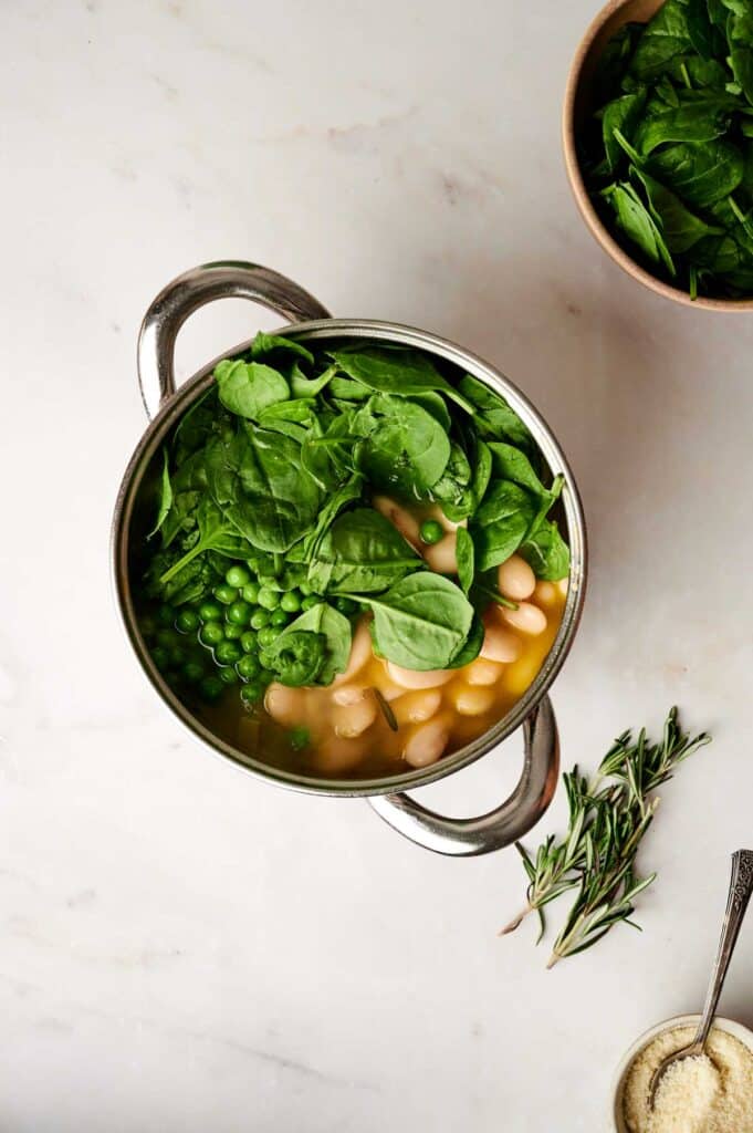 A pot of hearty vegetable soup brimming with fresh spinach, peas, and white beans in broth rests on a marble surface, accompanied by sprigs of rosemary and a bowl of greens nearby.