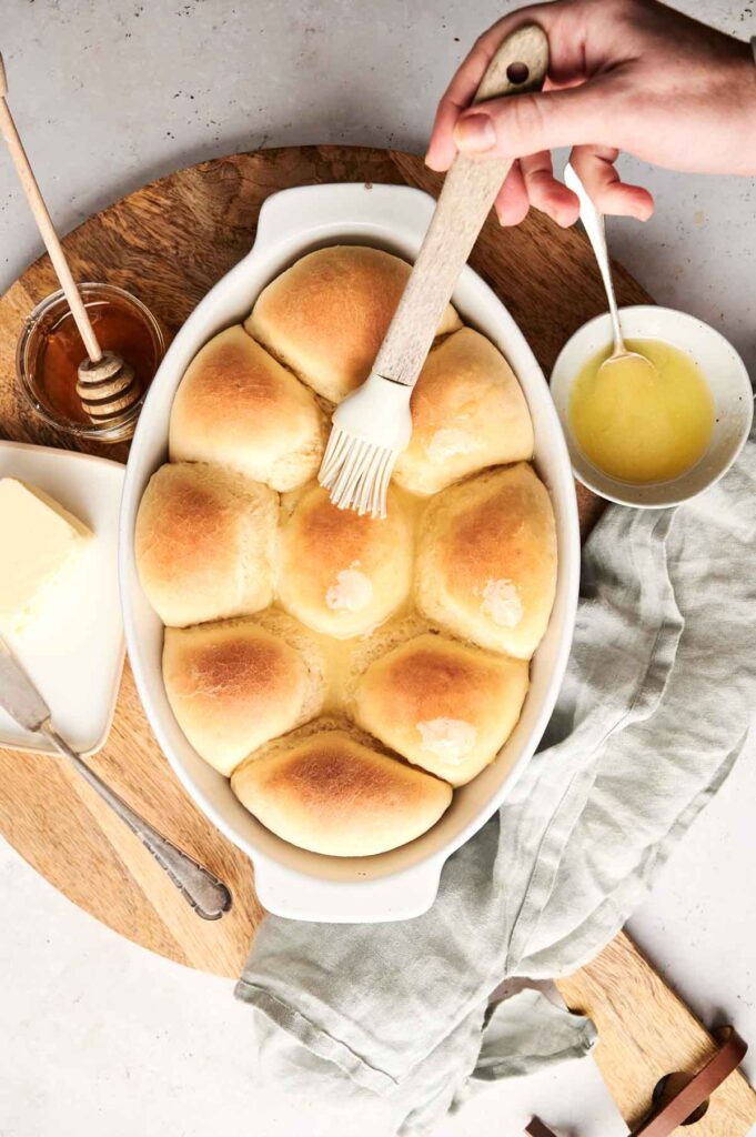 A hand brushes melted butter onto a pan of golden dinner rolls. Nearby are honey, a bowl with extra butter, and a folded cloth, completing the perfect dinner roll recipe scene.
