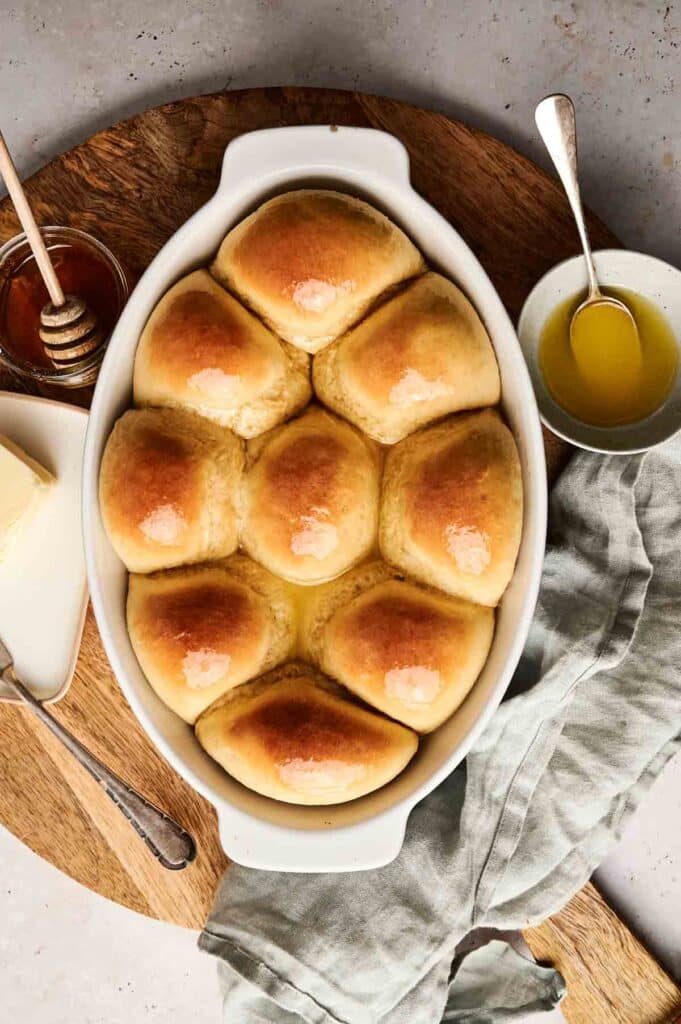 A baking dish with eight golden brown dinner rolls, fresh from a classic dinner roll recipe. A dish of butter and a bowl of honey are beside it on a wooden board. A silver spoon and a cloth napkin are also visible, ready to complete the perfect meal setting.