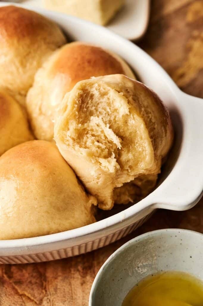 A warm, inviting dish of freshly baked bread rolls, with one partially torn to reveal its soft, airy texture alongside a small bowl of melted butter. Perfect for those seeking a simple yet delicious dinner roll recipe to elevate any meal.