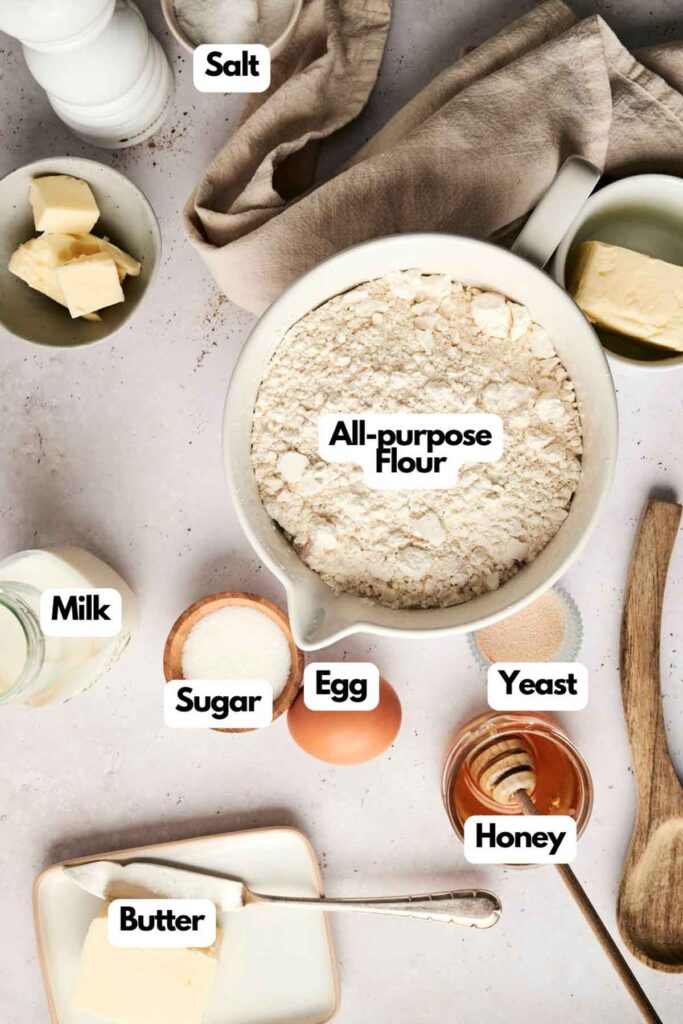 Ingredients for a delicious dinner roll recipe include flour, salt, butter, milk, sugar, an egg, yeast, and honey all neatly arranged on the countertop.
