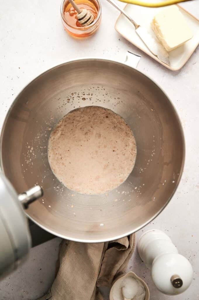 A metal mixing bowl with yeast mixture, surrounded by butter, a honey jar, salt shaker, and a cloth on the countertop&mdash;everything needed to start your dinner roll recipe.