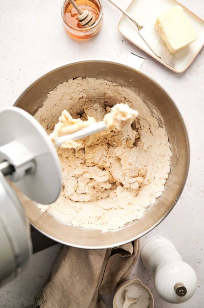 A mixer sits ready, surrounded by the essentials: dough, butter on a plate, a honey jar with a dipper, and a salt shaker on a light surface. Perfect ingredients for crafting your favorite dinner roll recipe.
