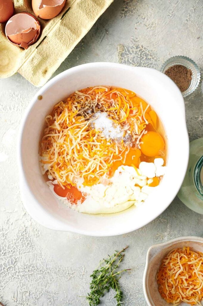 Overhead shot of a mixing bowl with cracked eggs, shredded cheese, sour cream, and seasonings ready to be whisked together.