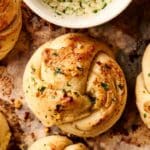 A close-up of garlic knots, perfectly golden and sprinkled with herbs, rests invitingly on a baking sheet, joined by a bowl of rich garlic butter dip.