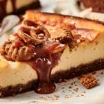 A slice of cheesecake topped with pecans, pretzels, and caramel sauce on a plate.